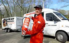 Ein Mann in rotem Anzug mit einem Feuerlöscher in der Hand vor einem Auto mit Anhänger