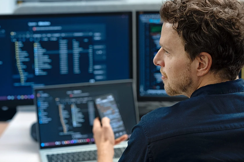 Ein Mann sitzt vor mehreren Bildschirmen mit Programmcode und blickt auf ein Mobiltelefon in seiner Hand.