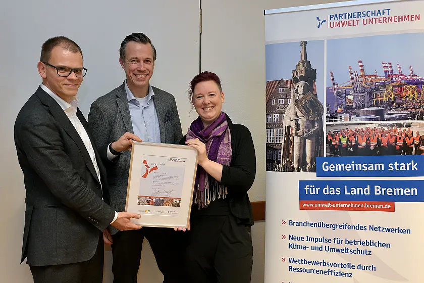 Jan-Hendrik Hunke und Heike Lau von der Bühnen GmbH & Co. KG nehmen die Auszeichnungsurkunde zum Klimaschutzbetrieb von Jan Fries, Staatsrat bei der Senatorin für Umwelt, Klima und Wissenschaft (links im Bild), entgegen.