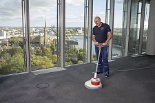 Ein Mann säubert mit einem Reinigungsgerät den Boden in einem modernen Bürogebäude. Im Hintergrund ist eine Stadtansicht zu erkennen.